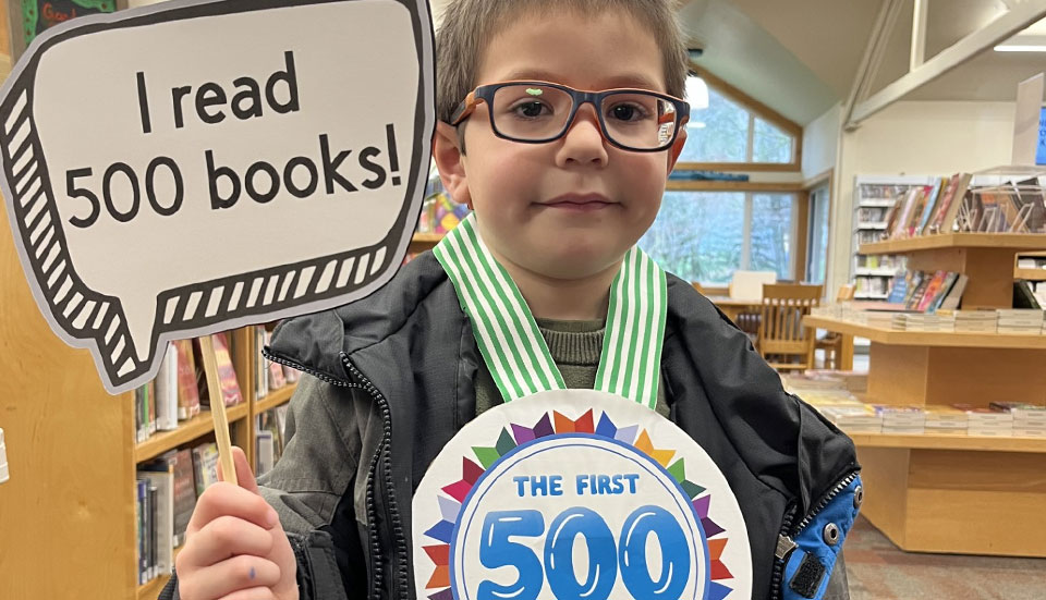young boy holding sign that says "I read 500 books" and wearing a large medal that says "The First 500"